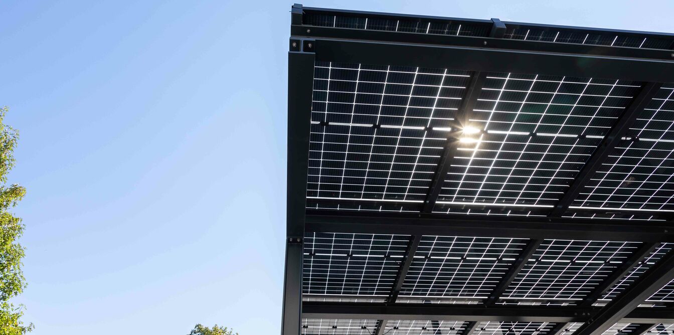 Ein Teil eines Solardachs aus mehreren Solarpaneelen, die in einem Metallrahmen montiert sind. Der Himmel ist klar und blau, und im Hintergrund sind Bäume zu sehen. Ein Teil eines Solardachs aus mehreren Solarpaneelen, die in einem Metallrahmen montiert sind. Der Himmel ist klar und blau, und im Hintergrund sind Bäume zu sehen.