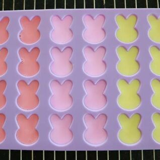 Eine lila Silikonform mit Hasenformen, die mit buntem Seifenmaterial gefüllt ist. Die Farben umfassen Rosa, Rot und Gelb und trocknen auf einem Rost. Eine lila Silikonform mit Hasenformen, die mit buntem Seifenmaterial gefüllt ist. Die Farben umfassen Rosa, Rot und Gelb und trocknen auf einem Rost.