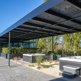 Ein moderner Außenbereich mit zwei Whirlpools unter einer Überdachung aus Solarpanelen. Umgeben von Pflanzen und Bäumen, mit einer gepflasterten Fläche Ein moderner Außenbereich mit zwei Whirlpools unter einer Überdachung aus Solarpanelen. Umgeben von Pflanzen und Bäumen, mit einer gepflasterten Fläche