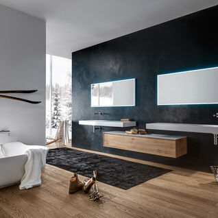 Badezimmer mit freistehender Badewanne, beleuchteten Spiegeln, Holzwaschtisch, Skiern an der Wand und großem Fenster. Elegante, gemütliche Atmosphäre.