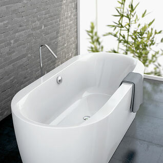 Eine freistehende, weiße Badewanne mit ergonomischem Design, vor einer grauen Steinwand. Große Fenster mit Blick auf Bambuspflanzen sorgen für ein natürliches Ambiente. Eine freistehende, weiße Badewanne mit ergonomischem Design, vor einer grauen Steinwand. Große Fenster mit Blick auf Bambuspflanzen sorgen für ein natürliches Ambiente.