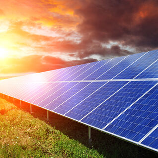 Eine Reihe von Solarpaneelen steht auf einem grünen Feld, während die Sonne am Horizont untergeht und den Himmel in warmen Farben erstrahlen lässt. Eine Reihe von Solarpaneelen steht auf einem grünen Feld, während die Sonne am Horizont untergeht und den Himmel in warmen Farben erstrahlen lässt.