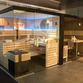 Ein moderner Saunaraum aus hellem Holz mit Glaswänden. Die Beleuchtung ist blau. Innen befinden sich Bänke, ein Holzeimer und ein Ofen mit Steinen. Ein moderner Saunaraum aus hellem Holz mit Glaswänden. Die Beleuchtung ist blau. Innen befinden sich Bänke, ein Holzeimer und ein Ofen mit Steinen.