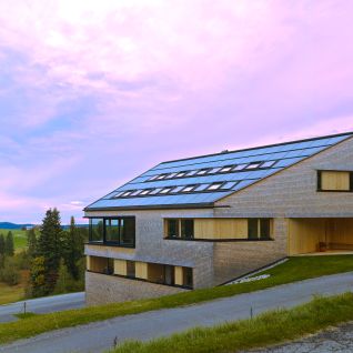 Modernes Haus mit geneigtem Dach und Solarpanelen, umgeben von gr&uuml;nen Wiesen und B&auml;umen. Der Himmel zeigt ein beeindruckendes rosa-blaues Farbspiel.