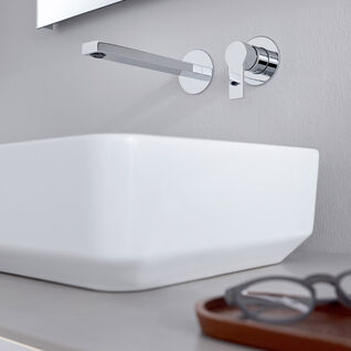 Ein elegantes, modernes Waschbecken mit Wandarmatur. Auf der Ablage liegt eine Brille in einem Tablett. Schlichtes und funktionales Design.