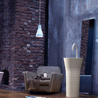 Ein freistehendes, kunstvolles Waschbecken in einem industriellen Loft mit Backsteinwänden, Sessel und Hängelampe im Hintergrund. Ein freistehendes, kunstvolles Waschbecken in einem industriellen Loft mit Backsteinwänden, Sessel und Hängelampe im Hintergrund.