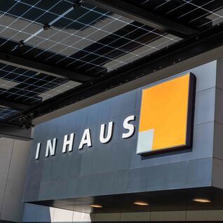 Ein Gebäude mit einem Solardach, das aus mehreren Solarpaneelen besteht. An der Fassade des Gebäudes befindet sich ein Schild mit der Aufschrift „INHAUS“ und einem orangefarbenen Logo. Solarcarport