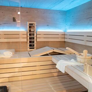 Ein moderner Saunaraum aus hellem Holz, mit Glaswänden und blauer Beleuchtung. Bänke mit Sauna-Zubehör und ein Ofen mit Steinen sind sichtbar. Ein moderner Saunaraum aus hellem Holz, mit Glaswänden und blauer Beleuchtung. Bänke mit Sauna-Zubehör und ein Ofen mit Steinen sind sichtbar.