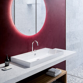 Modernes Badezimmer mit auff&auml;lliger roter Wand, rundem Spiegel mit Hintergrundbeleuchtung, schlichtem wei&szlig;en Waschbecken und einem Holzregal darunter. Stilvoll und elegant.