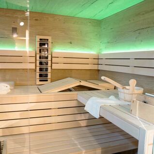 Ein moderner Saunaraum aus hellem Holz mit einer Glasfront. Die Beleuchtung ist in einem beruhigenden Grün gehalten. Auf den Bänken liegen Sauna-Zubehör wie ein Holzeimer und ein Handtuch. Ein moderner Saunaraum aus hellem Holz mit einer Glasfront. Die Beleuchtung ist in einem beruhigenden Grün gehalten. Auf den Bänken liegen Sauna-Zubehör wie ein Holzeimer und ein Handtuch.