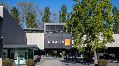 Modernes Gebäude "INHAUS" mit großen Fenstern und geradliniger Architektur. Ein Baum und Pflanzen zieren den Vorplatz unter klarem, blauem Himmel.