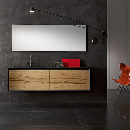 Modernes Badezimmer mit dunkler Wand, h&auml;ngendem Holzschrank, gro&szlig;em Spiegel, stilvoller Beleuchtung, schwarzem Boden, orangefarbenem Stuhl und kleinem Tisch.