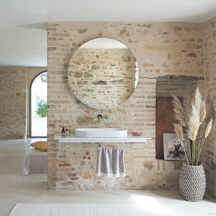 Ein Badezimmer mit rustikaler Backsteinwand und rundem Spiegel. Ein moderner Waschtisch, Dekorationen wie ein Korb mit Pampasgras und ein helles, einladendes Ambiente runden den Raum ab. Ein Badezimmer mit rustikaler Backsteinwand und rundem Spiegel. Ein moderner Waschtisch, Dekorationen wie ein Korb mit Pampasgras und ein helles, einladendes Ambiente runden den Raum ab.