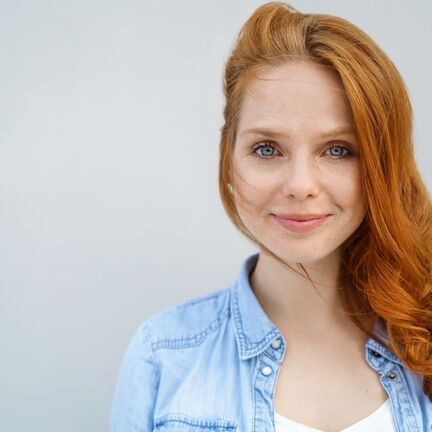 Eine lächelnde Person mit roten Haaren trägt ein helles Jeanshemd und steht vor einem schlichten, hellen Hintergrund. Eine lächelnde Person mit roten Haaren trägt ein helles Jeanshemd und steht vor einem schlichten, hellen Hintergrund.
