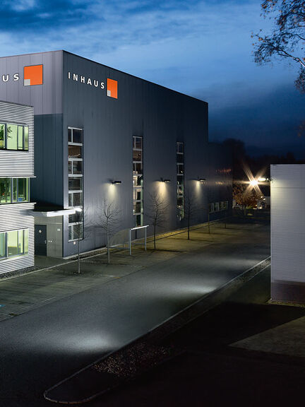 Ein modernes Bürogebäude bei Nacht, beleuchtet von innen. Es hat drei Etagen, große Fenster und eine gepflegte Umgebung mit Bäumen und Gehwegen. Ein modernes Bürogebäude bei Nacht, beleuchtet von innen. Es hat drei Etagen, große Fenster und eine gepflegte Umgebung mit Bäumen und Gehwegen.