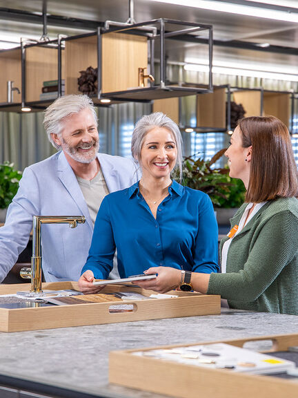 Drei Personen stehen an einem Küchenausstellungsbereich. Eine Frau zeigt einem Paar verschiedene Materialproben oder Farbmuster.  Drei Personen stehen an einem Küchenausstellungsbereich. Eine Frau zeigt einem Paar verschiedene Materialproben oder Farbmuster.