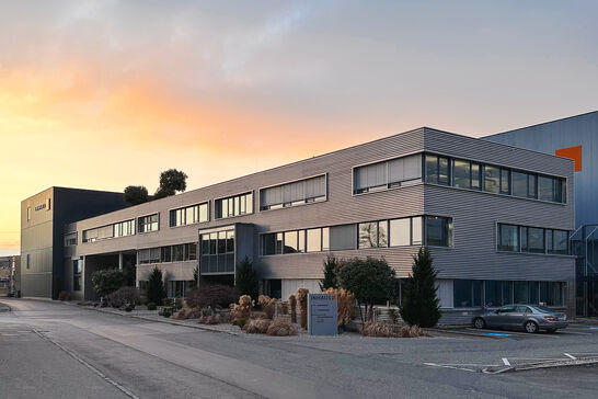 Modernes Bürogebäude mit großen Fenstern, umgeben von gepflegter Landschaftsgestaltung. Der Himmel im Hintergrund zeigt einen schönen Sonnenuntergang. Modernes Bürogebäude mit großen Fenstern, umgeben von gepflegter Landschaftsgestaltung. Der Himmel im Hintergrund zeigt einen schönen Sonnenuntergang.