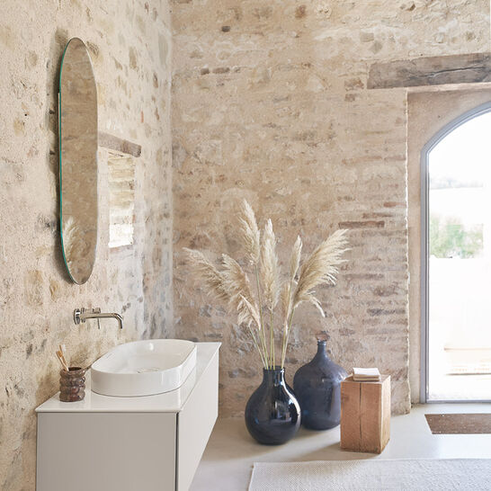 Rustikales Badezimmer mit Natursteinwänden und ovalem Spiegel. Moderner, hängender Waschtisch und dekorative Vasen mit Pampasgras. Große Tür mit Blick nach draußen. Rustikales Badezimmer mit Natursteinwänden und ovalem Spiegel. Moderner, hängender Waschtisch und dekorative Vasen mit Pampasgras. Große Tür mit Blick nach draußen.