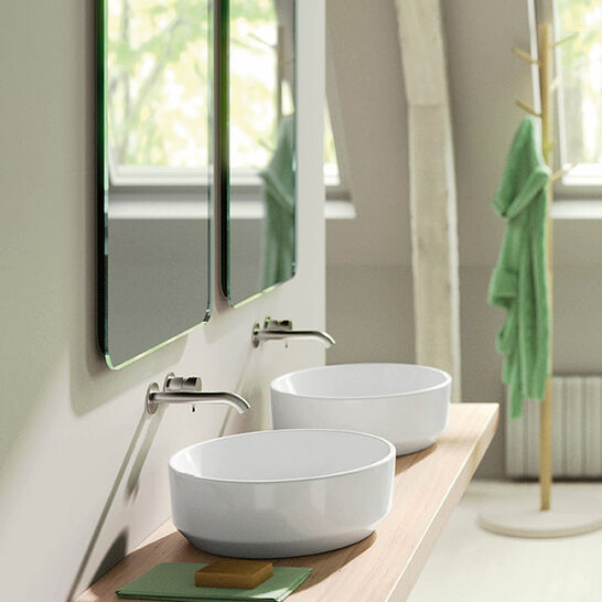 Ein stilvolles Badezimmer mit zwei runden Aufsatzwaschbecken auf einem Holzbrett. Darüber befinden sich zwei Spiegel. Im Hintergrund hängt ein grünes Handtuch. Modern und elegant gestaltet.