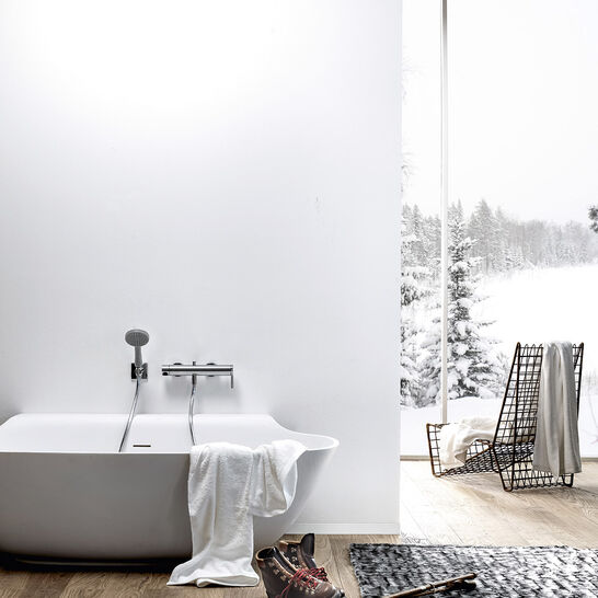 Helles Badezimmer mit freistehender Badewanne, großem Fenster mit Blick auf verschneite Landschaft, Holzfußboden und minimalistischer Dekoration. Skistock und Schuhe als Akzent.