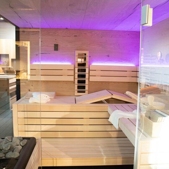 Ein moderner Saunaraum aus hellem Holz, mit Glaswänden und lila Beleuchtung. Bänke mit Sauna-Zubehör und ein Ofen mit Steinen sind sichtbar. Ein moderner Saunaraum aus hellem Holz, mit Glaswänden und lila Beleuchtung. Bänke mit Sauna-Zubehör und ein Ofen mit Steinen sind sichtbar.