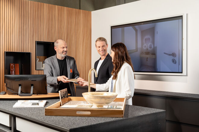 Ein Verkäufer berät ein Paar in einem modernen Ausstellungsraum. Sie lachen und tauschen Dokumente aus, während sie sich Badezimmerprodukte ansehen. Ein Verkäufer berät ein Paar in einem modernen Ausstellungsraum. Sie lachen und tauschen Dokumente aus, während sie sich Badezimmerprodukte ansehen.