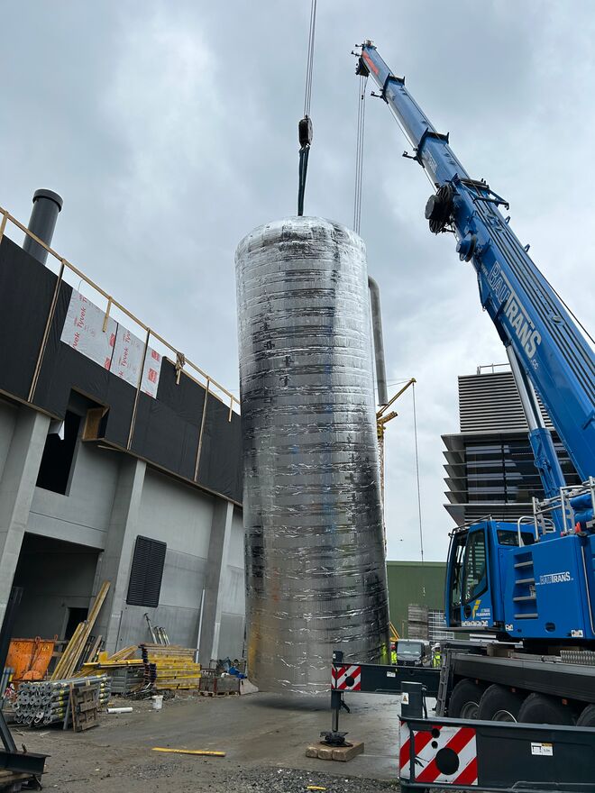 Ein blauer Kran hebt einen großen, silbernen Zylinder vor einem Industriegebäude. Der Himmel ist bewölkt, die Szene wirkt industriell. Ein blauer Kran hebt einen großen, silbernen Zylinder vor einem Industriegebäude. Der Himmel ist bewölkt, die Szene wirkt industriell.