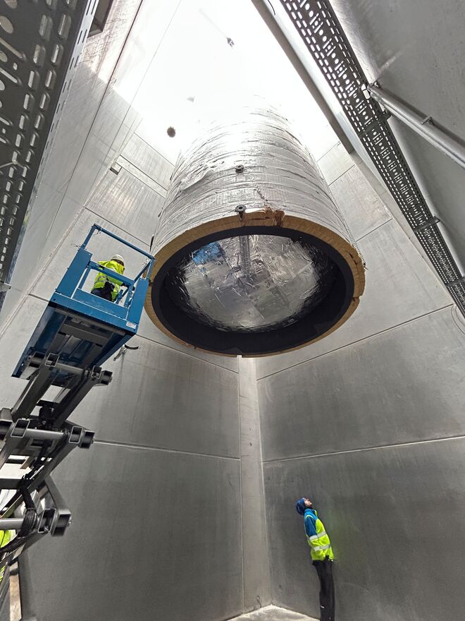 Ein großer Zylinder wird in einen Schacht abgesenkt. Ein Arbeiter steht auf einer Hebebühne, ein anderer beobachtet von unten. Die Umgebung ist industriell. Ein großer Zylinder wird in einen Schacht abgesenkt. Ein Arbeiter steht auf einer Hebebühne, ein anderer beobachtet von unten. Die Umgebung ist industriell.