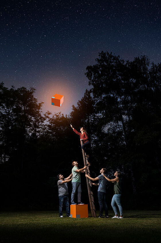 Eine Gruppe von Menschen hält eine Leiter, während eine Person versucht, einen leuchtenden orangefarbenen Würfel zu erreichen, unter einem sternenklaren Himmel. Eine Gruppe von Menschen hält eine Leiter, während eine Person versucht, einen leuchtenden orangefarbenen Würfel zu erreichen, unter einem sternenklaren Himmel.