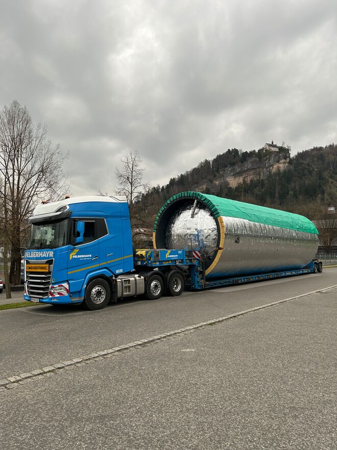 Ein blauer LKW transportiert einen großen, silbernen Zylinder auf einem Anhänger. Der Himmel ist bewölkt, im Hintergrund sind Hügel zu sehen. Ein blauer LKW transportiert einen großen, silbernen Zylinder auf einem Anhänger. Der Himmel ist bewölkt, im Hintergrund sind Hügel zu sehen.
