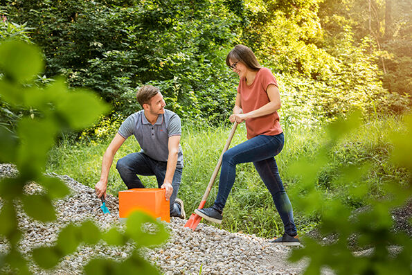 Zwei Personen arbeiten auf einem Kiesweg in einer grünen Umgebung. Eine Person kniet mit einem orangefarbenen Eimer, die andere steht mit einem Besen. Zwei Personen arbeiten auf einem Kiesweg in einer grünen Umgebung. Eine Person kniet mit einem orangefarbenen Eimer, die andere steht mit einem Besen.