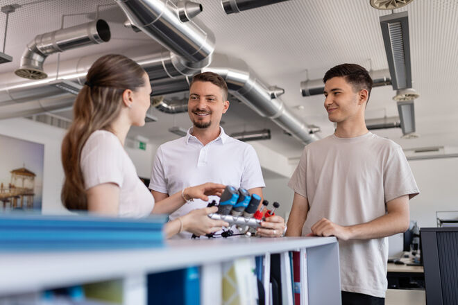 Drei Personen stehen in einem modernen Büro mit Lüftungsrohren und sprechen. Zwei Männer und eine Frau halten kleine Modelle in der Hand. Drei Personen stehen in einem modernen Büro mit Lüftungsrohren und sprechen. Zwei Männer und eine Frau halten kleine Modelle in der Hand.