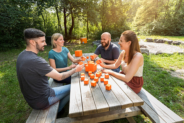 Vier Personen sitzen an einem Picknicktisch im Freien und spielen mit orangefarbenen Würfeln. Sie wirken fröhlich und entspannt. Vier Personen sitzen an einem Picknicktisch im Freien und spielen mit orangefarbenen Würfeln. Sie wirken fröhlich und entspannt.