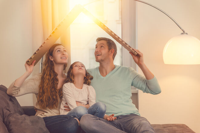 Eine Familie sitzt auf einem Sofa und hält ein aus Pappe geformtes Dreieck über sich, das ein Dach symbolisiert. Sie blicken lächelnd nach oben in das warme Licht einer Stehlampe. Eine Familie sitzt auf einem Sofa und hält ein aus Pappe geformtes Dreieck über sich, das ein Dach symbolisiert. Sie blicken lächelnd nach oben in das warme Licht einer Stehlampe.