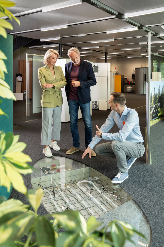 Drei Personen befinden sich in einem modernen Raum. Ein Mann kniet und zeigt auf den Boden, während das Paar interessiert zusieht. Pflanzen sind im Vordergrund. Drei Personen befinden sich in einem modernen Raum. Ein Mann kniet und zeigt auf den Boden, während das Paar interessiert zusieht. Pflanzen sind im Vordergrund.