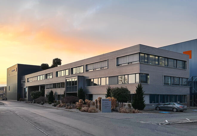 Modernes Bürogebäude mit großen Fenstern, umgeben von gepflegter Landschaftsgestaltung. Der Himmel im Hintergrund zeigt einen schönen Sonnenuntergang. Modernes Bürogebäude mit großen Fenstern, umgeben von gepflegter Landschaftsgestaltung. Der Himmel im Hintergrund zeigt einen schönen Sonnenuntergang.