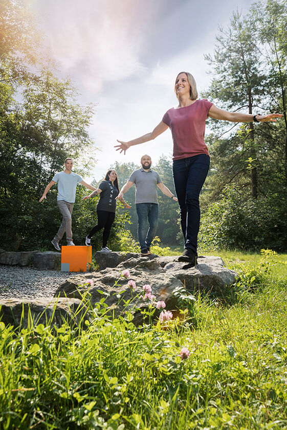 Vier Personen balancieren mit ausgebreiteten Armen auf Steinen in einem sonnigen, grünen Park. Sie wirken fröhlich und entspannt. Vier Personen balancieren mit ausgebreiteten Armen auf Steinen in einem sonnigen, grünen Park. Sie wirken fröhlich und entspannt.