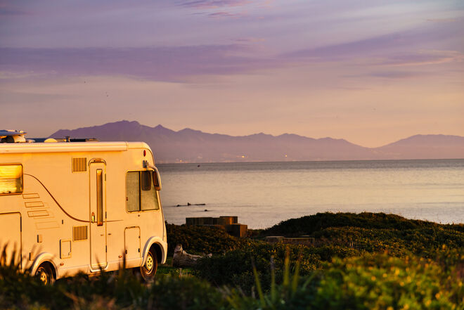 Ein Wohnmobil steht in einer malerischen Küstenlandschaft bei Sonnenuntergang. Im Hintergrund sind das Meer und Berge zu sehen, während der Himmel in warmen Farben leuchtet. Ein Wohnmobil steht in einer malerischen Küstenlandschaft bei Sonnenuntergang. Im Hintergrund sind das Meer und Berge zu sehen, während der Himmel in warmen Farben leuchtet.