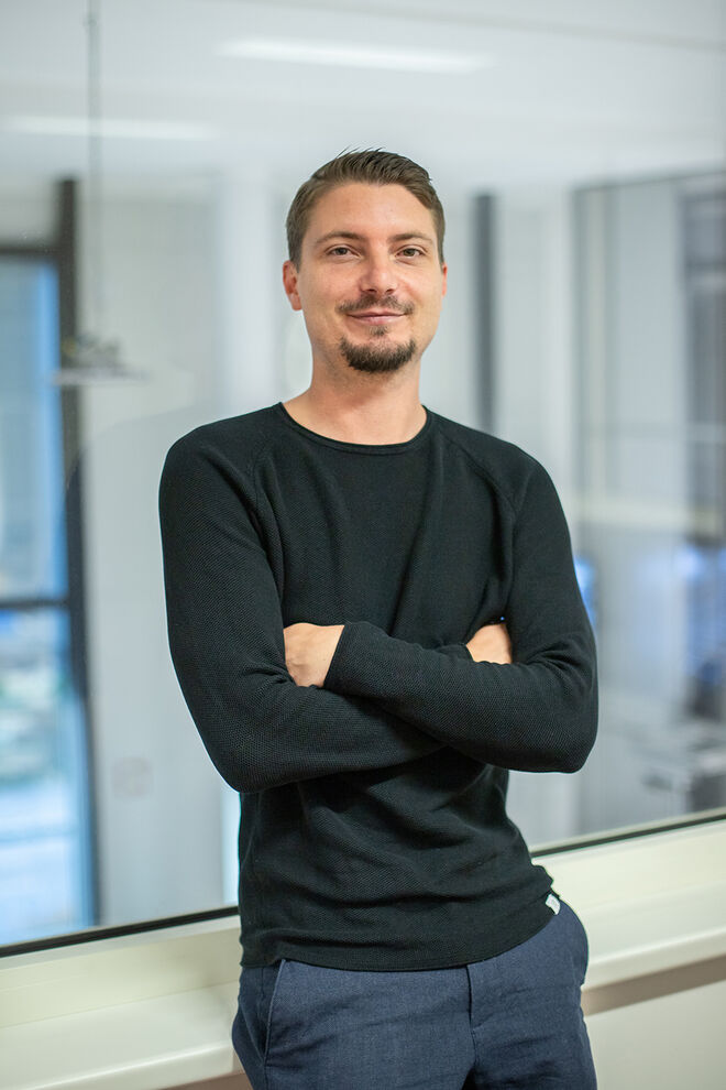 Ein Mann mit kurzem Haar steht lächelnd in einem modernen Büro, die Arme verschränkt. Er trägt einen schwarzen Pullover und blaue Hose. Ein Mann mit kurzem Haar steht lächelnd in einem modernen Büro, die Arme verschränkt. Er trägt einen schwarzen Pullover und blaue Hose.