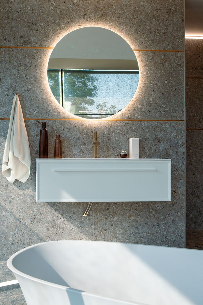 Modernes Badezimmer mit beleuchtetem Spiegel, grauer Steinwand, weißem Waschtisch und freistehender Wanne. Modernes Badezimmer mit beleuchtetem Spiegel, grauer Steinwand, weißem Waschtisch und freistehender Wanne.