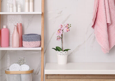 Ein stilvoll eingerichtetes Badezimmerregal mit rosa und grauen Accessoires. Eine Orchidee in einem dekorativen Topf steht auf einem weißen Tisch. Ein rosa Handtuch hängt daneben. Ein stilvoll eingerichtetes Badezimmerregal mit rosa und grauen Accessoires. Eine Orchidee in einem dekorativen Topf steht auf einem weißen Tisch. Ein rosa Handtuch hängt daneben.