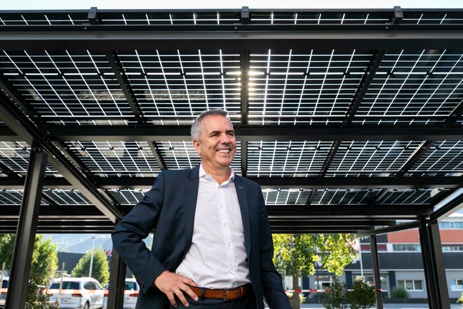 Ein Mann in einem Anzug steht unter einem Solardach, das aus mehreren Solarpaneelen besteht. Er lächelt und hat eine Hand in der Hosentasche. Ein Mann in einem Anzug steht unter einem Solardach, das aus mehreren Solarpaneelen besteht. Er lächelt und hat eine Hand in der Hosentasche.