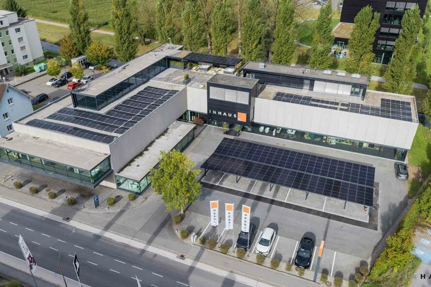 Luftbild eines U-förmigen Gebäudekomplexes "INHAUS" mit Solarpaneelen. Von Bäumen umgeben, Parkplatz und Flaggen entlang der Straße. Luftbild eines U-förmigen Gebäudekomplexes "INHAUS" mit Solarpaneelen. Von Bäumen umgeben, Parkplatz und Flaggen entlang der Straße.